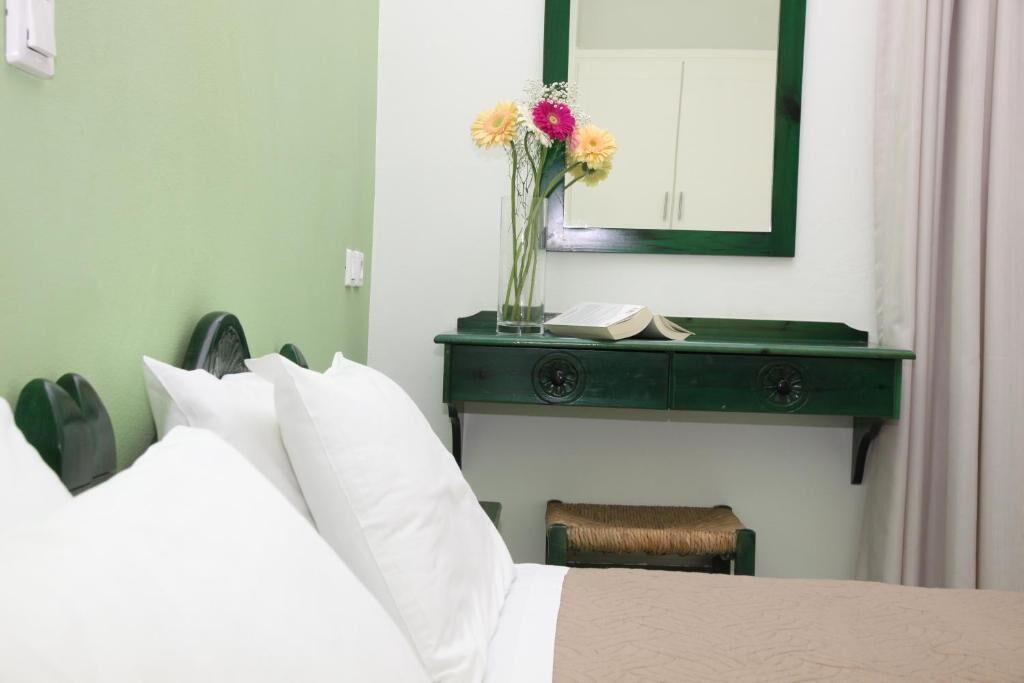 A hotel room with a bed, a green nightstand with a book, a vase with flowers, and a mirror on the wall.