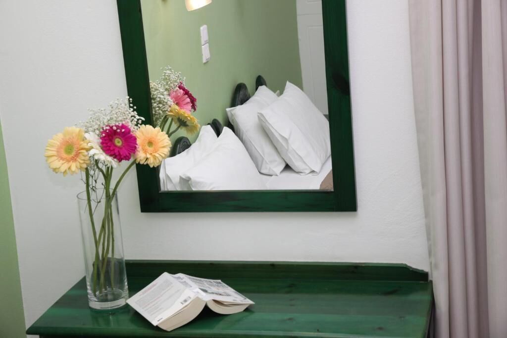Une pièce avec un lit, un miroir qui reflète le lit, un vase avec des fleurs et un livre posé sur une table verte.