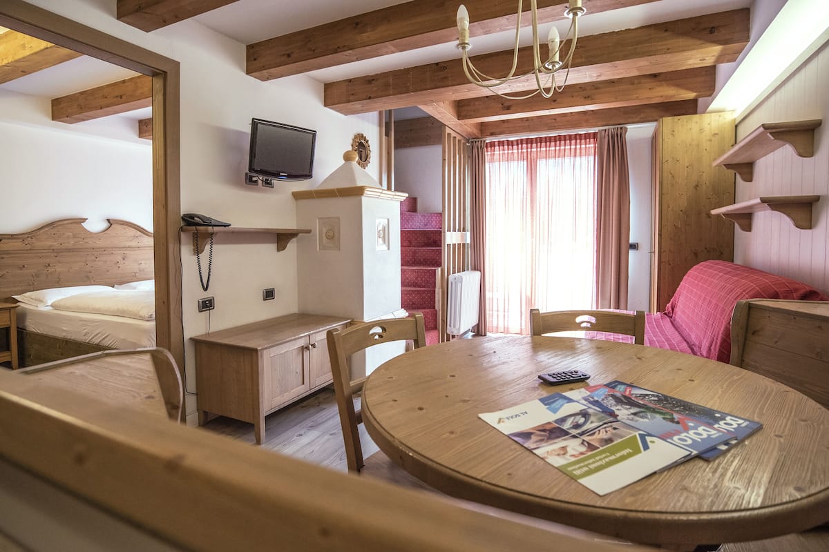 A cozy room with a wooden bed, a round dining table with magazines, a TV on the wall, and a staircase leading to an upper level.