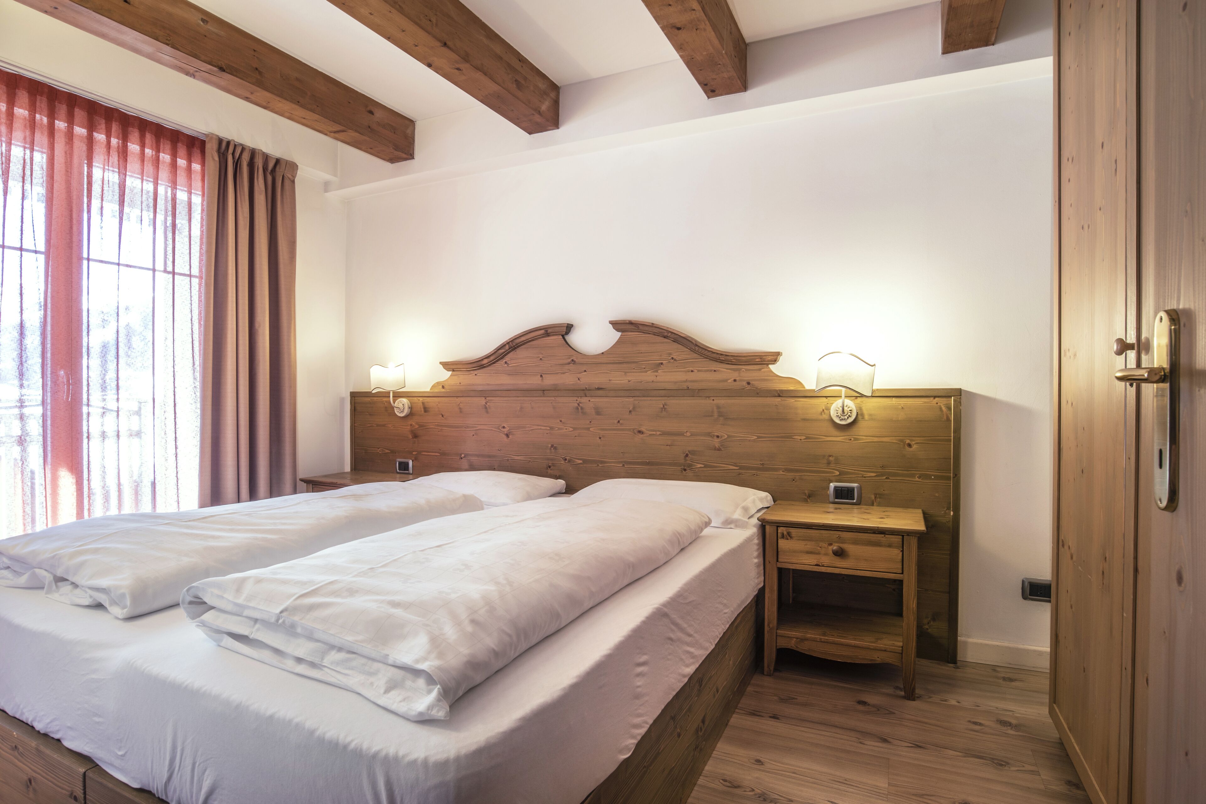 A double bed with white linens, a wooden headboard with wall-mounted lights, a nightstand with a drawer, and a wooden door.
