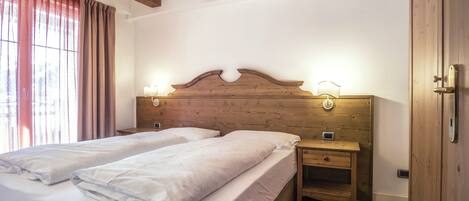 A double bed with white linens, a wooden headboard with wall-mounted lights, a nightstand with a drawer, and a wooden door.