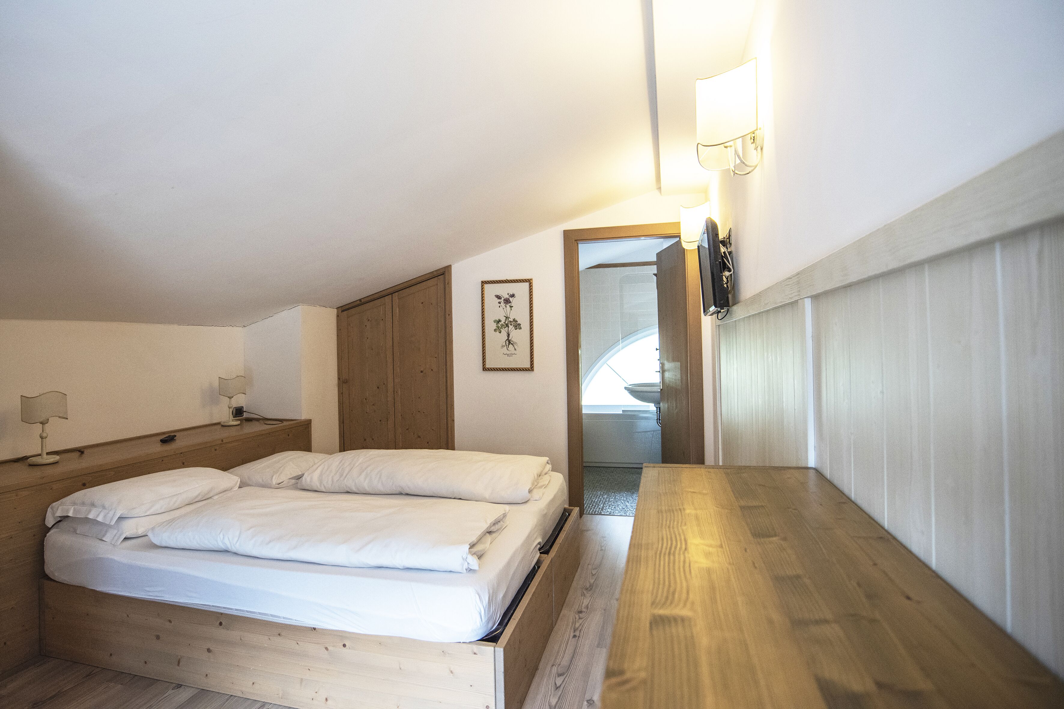 A bedroom with a wooden bed, bedside tables, and a nightstand. There is a framed picture on the wall and a mirror above the bed.