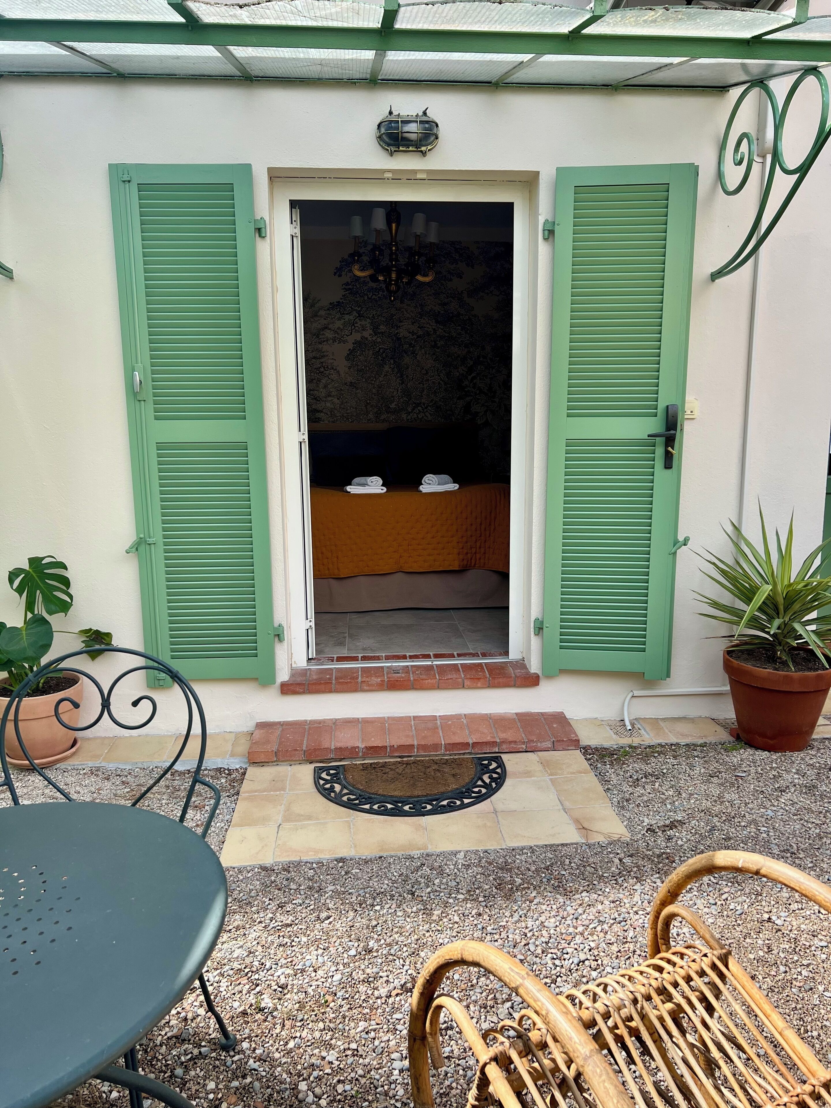 A hotel entrance with green shutters, a bed with towels, a potted plant, and a wicker chair.
