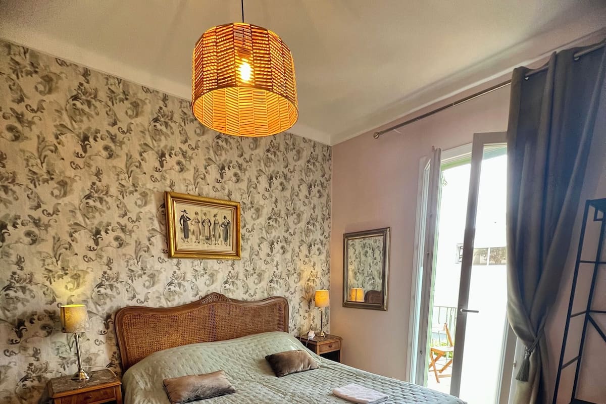 Ein Schlafzimmer mit einem Bett, zwei Nachttischen, einer Hängeleuchte, einem Fenster mit Vorhängen und einem Sessel.
