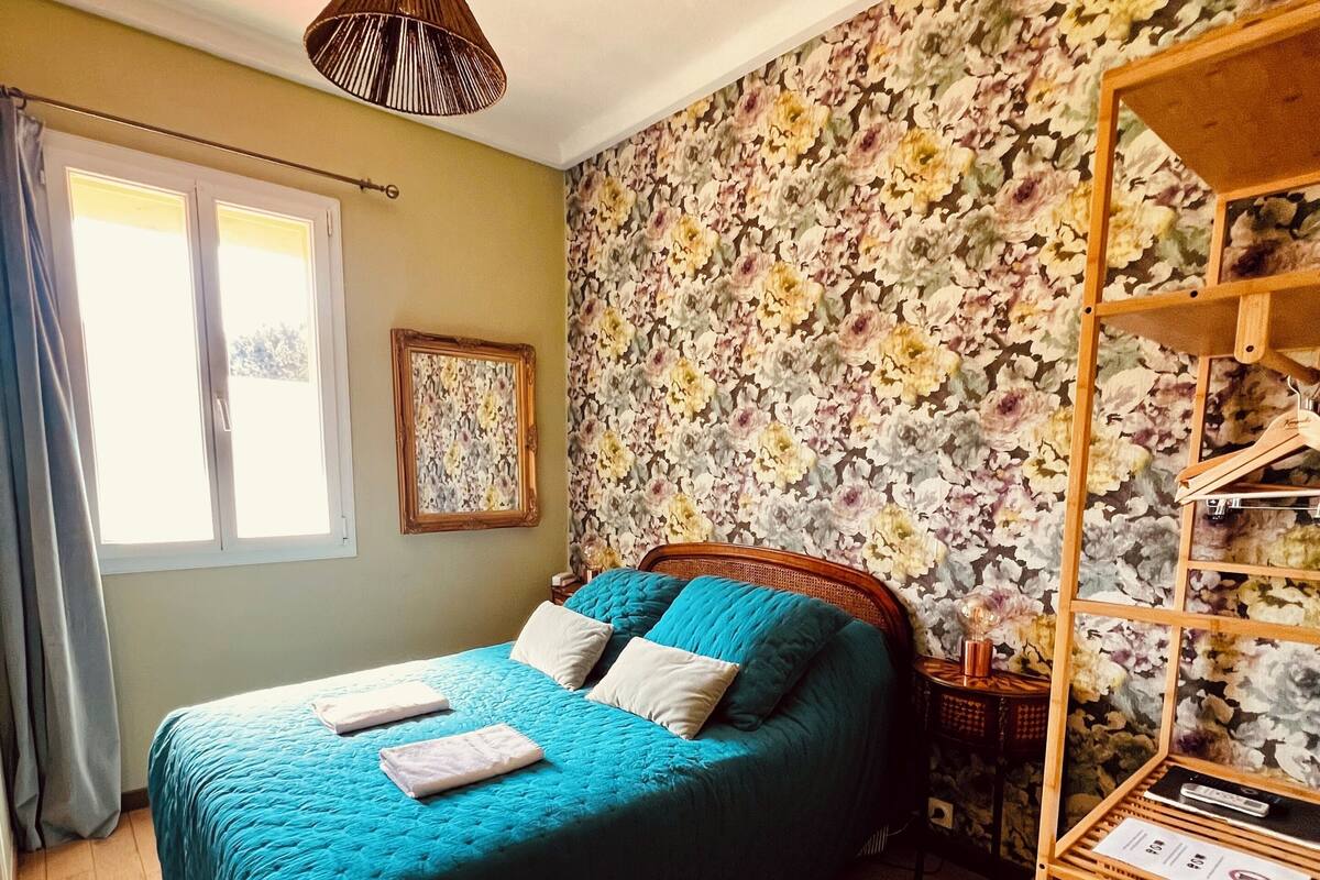 Ein Zimmer mit einem Bett, einem Holzstuhl, einer Pendelleuchte, floralem Wandpapier und einem Fenster mit Vorhängen.
