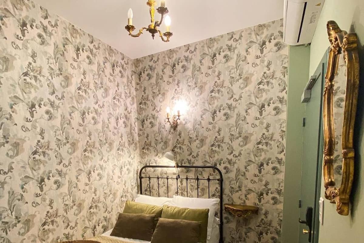 Ein Schlafzimmer mit Blumenwallpaper, einem Bett mit grünen und braunen Kissen, einem Nachttisch, einem Sessel, einem Hut, Sonnenbrillen und einer Chandelier.