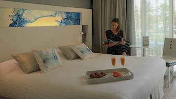 Une chambre d’hôtel avec un lit, un plateau contenant des croissants et de la pastèque, deux verres de jus d’orange et une femme qui lit un livre.