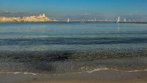 Un paysage côtier avec une plage de sable, une mer calme et, au loin, un promontoire rocheux sur lequel se trouve une construction.