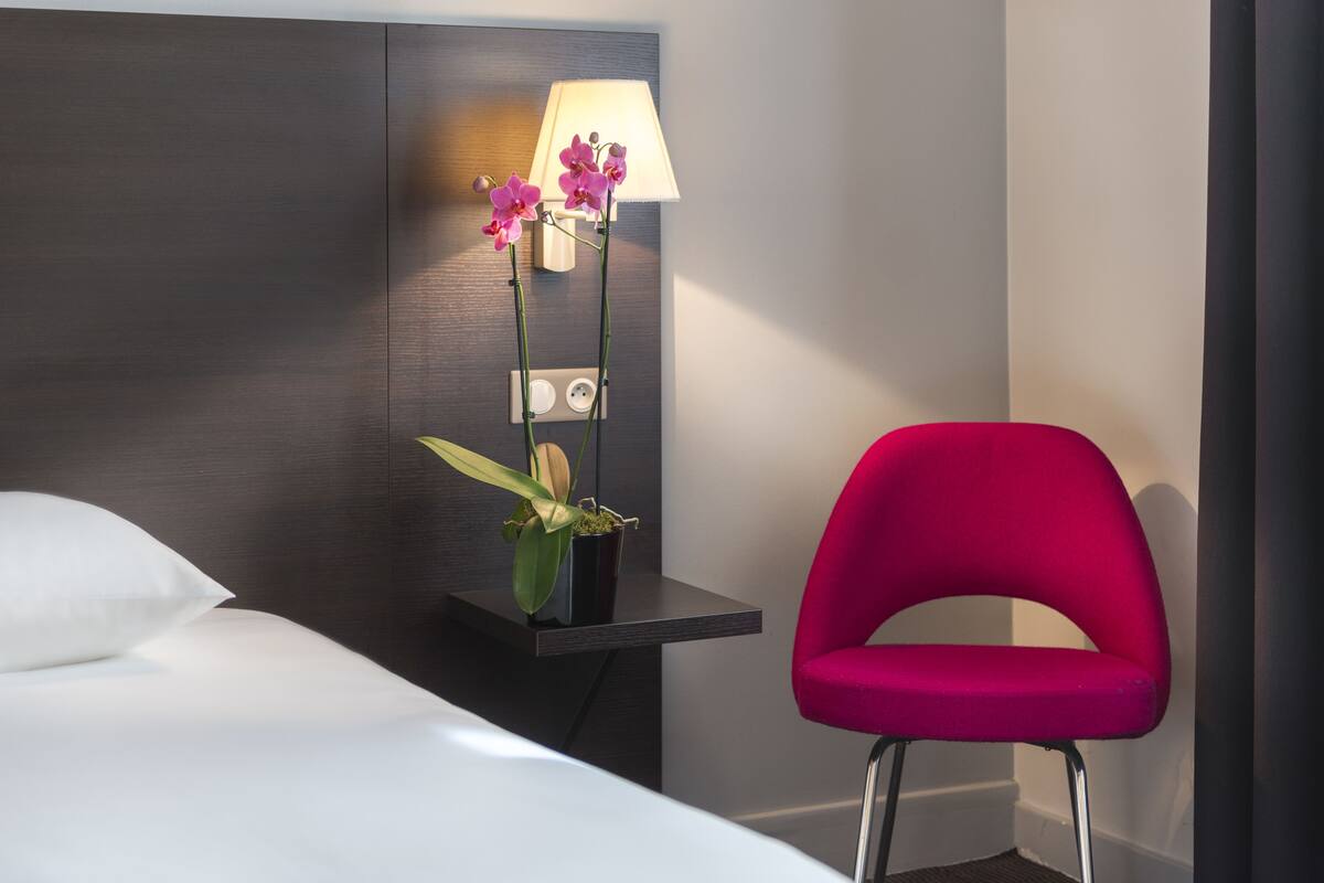 A hotel room with a bed, a nightstand with a lamp and orchids, and a bright red chair.