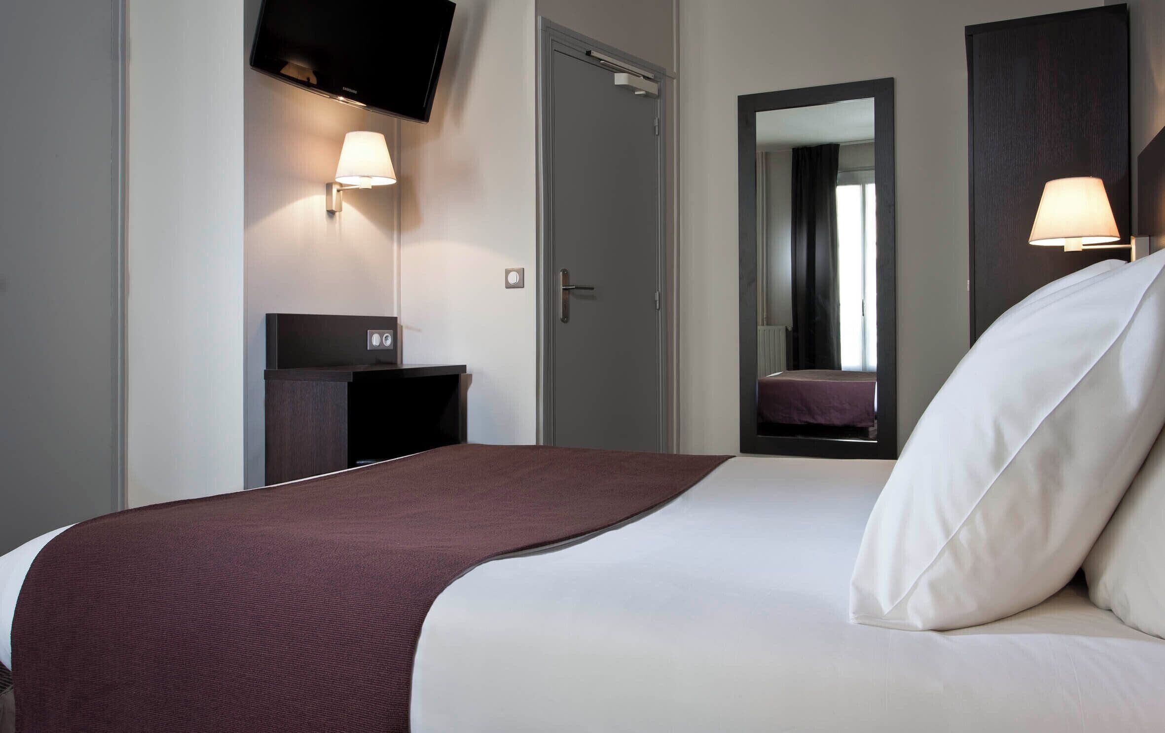 A hotel room with a bed, a TV mounted on the wall, a bedside table, and a mirror.