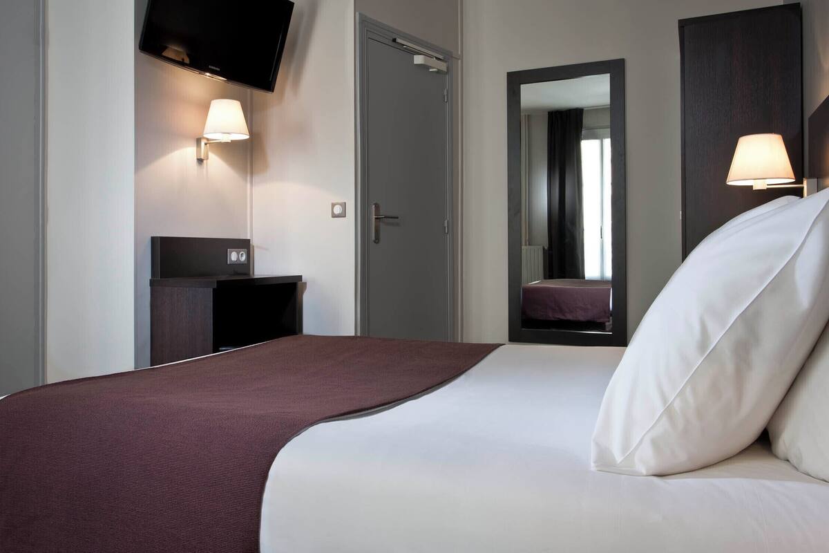 A hotel room with a bed, a TV mounted on the wall, a bedside table, and a mirror.