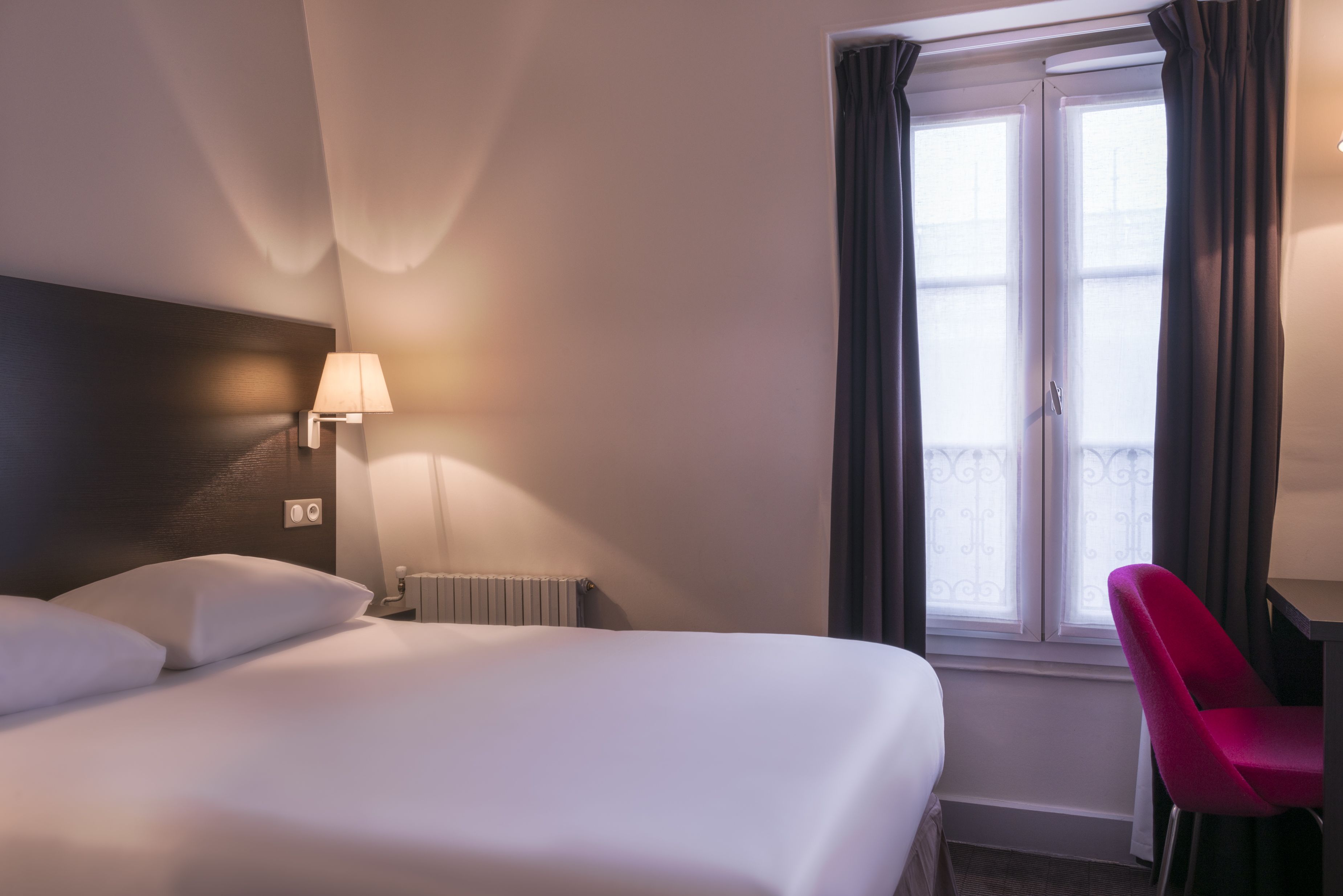 Habitación de hotel con cama, escritorio, silla, lámpara y ventana con cortinas.