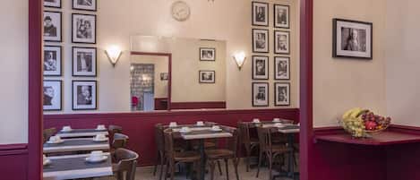 A restaurant with tables and chairs, framed pictures on the walls, and a fruit bowl on a counter.