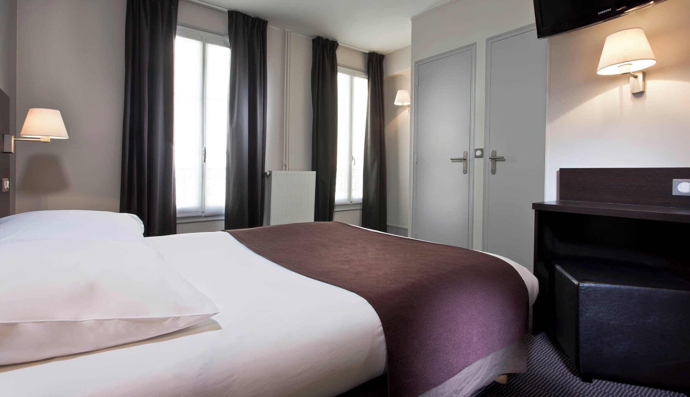 A hotel room with a bed, a desk, a chair, a wardrobe, and a lamp.