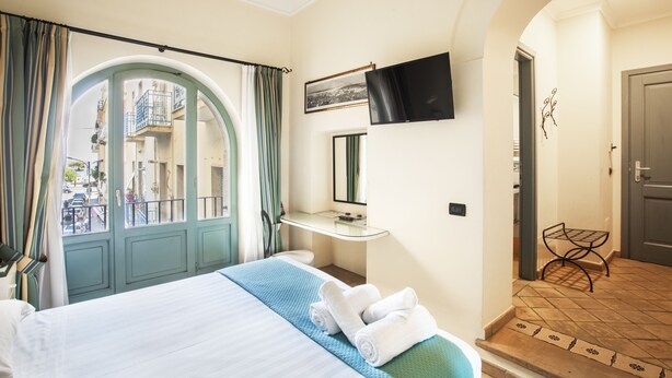A hotel room with a bed, a TV mounted on the wall, a bathroom with a mirror, and a view of the street through an arched door. - Quartuccio - Albergo Diffuso (Gaeta)