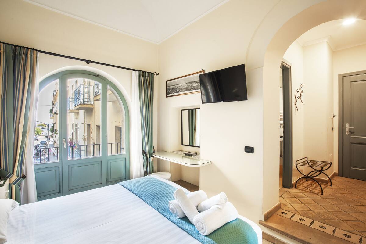 A hotel room with a bed, a TV mounted on the wall, a bathroom with a mirror, and a view of the street through an arched door.