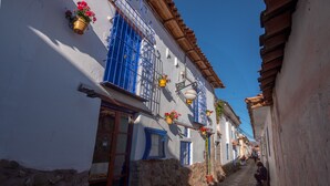 A narrow cobblestone street lined with white buildings featuring blue window frames and doors, stone walls, and hanging flower pots. - Casona les Pleiades (Cusco)