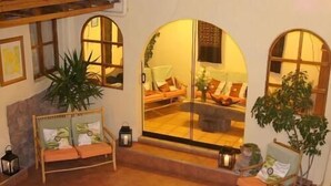 A cozy living room with a sofa, armchairs, and a coffee table. There are two large windows with arched tops, a fireplace, and several potted plants. - Casona les Pleiades (Cusco)