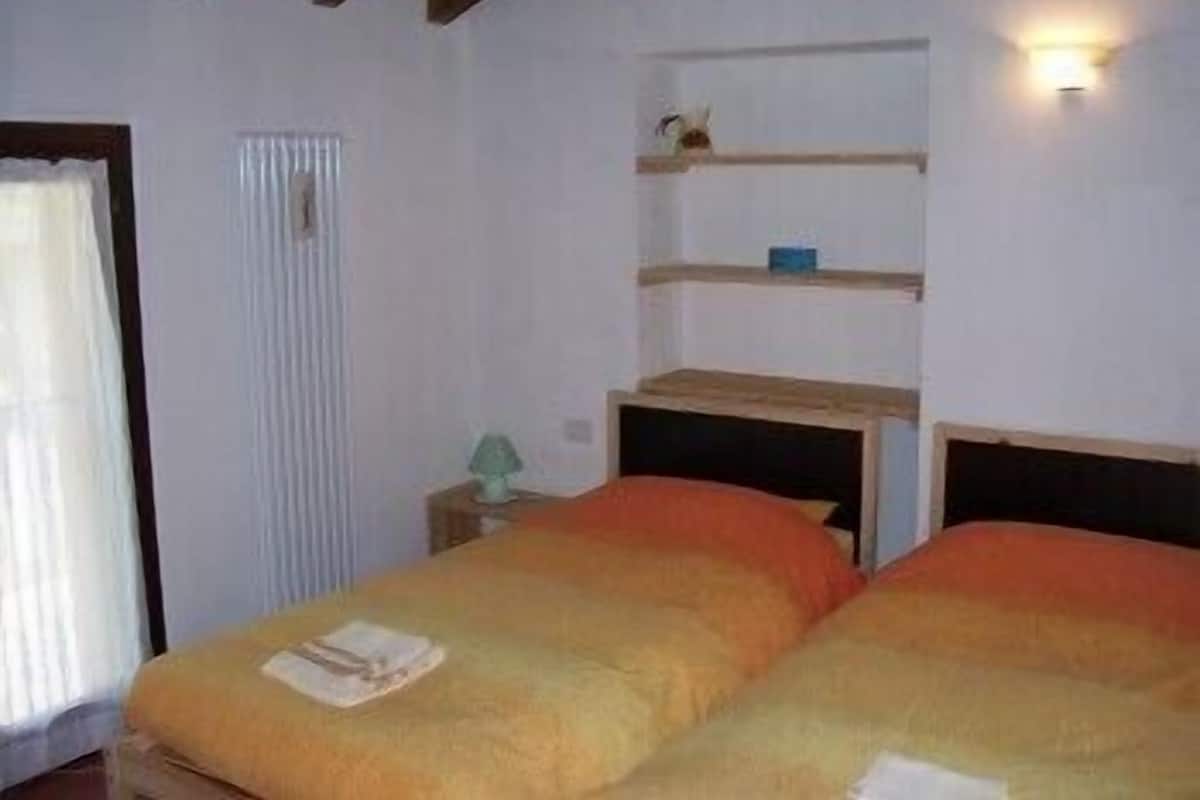 A room with two beds, a window with curtains, a shelf with a lamp, and a wall-mounted light.