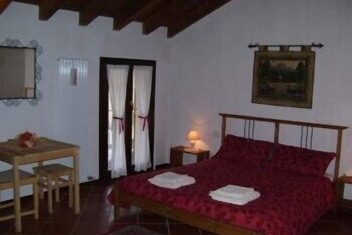 A bedroom with a wooden bed, a small table, a chair, a nightstand with a lamp, and a painting on the wall.