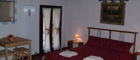 Un dormitorio con una cama de madera, una mesita pequeña, una silla, una mesita de noche con lámpara y un cuadro en la pared.
