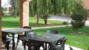 Un espace extérieur aménagé en terrasse, avec des piliers en briques, des tables et des chaises en plastique, et une vue sur une pelouse verdoyante et des arbres.