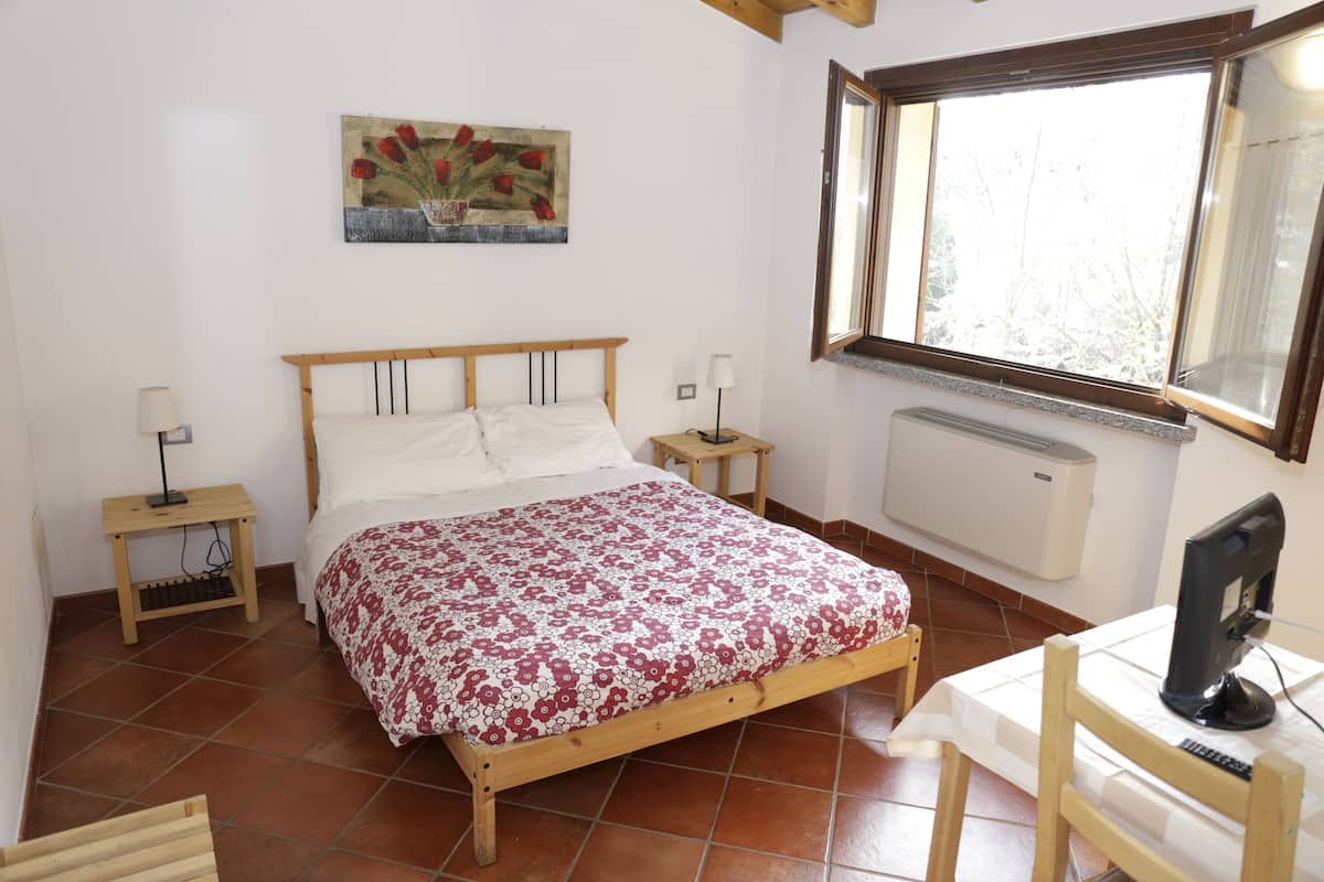 A bedroom with a wooden bed, two bedside tables, a desk with a computer, a window with a view, and a radiator.