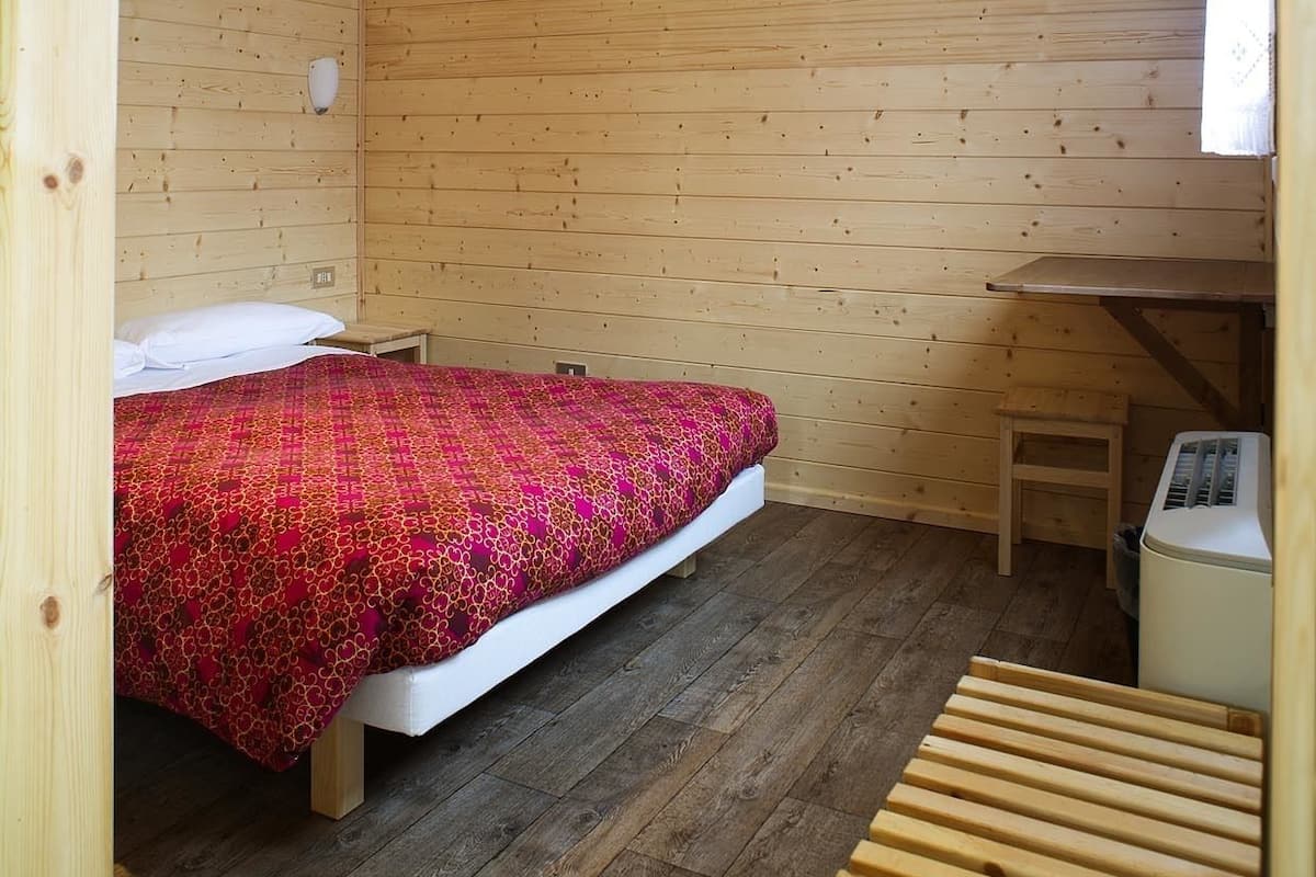 A small, cozy room with a wooden floor and walls, a bed with a patterned blanket, a small table, and a heater.