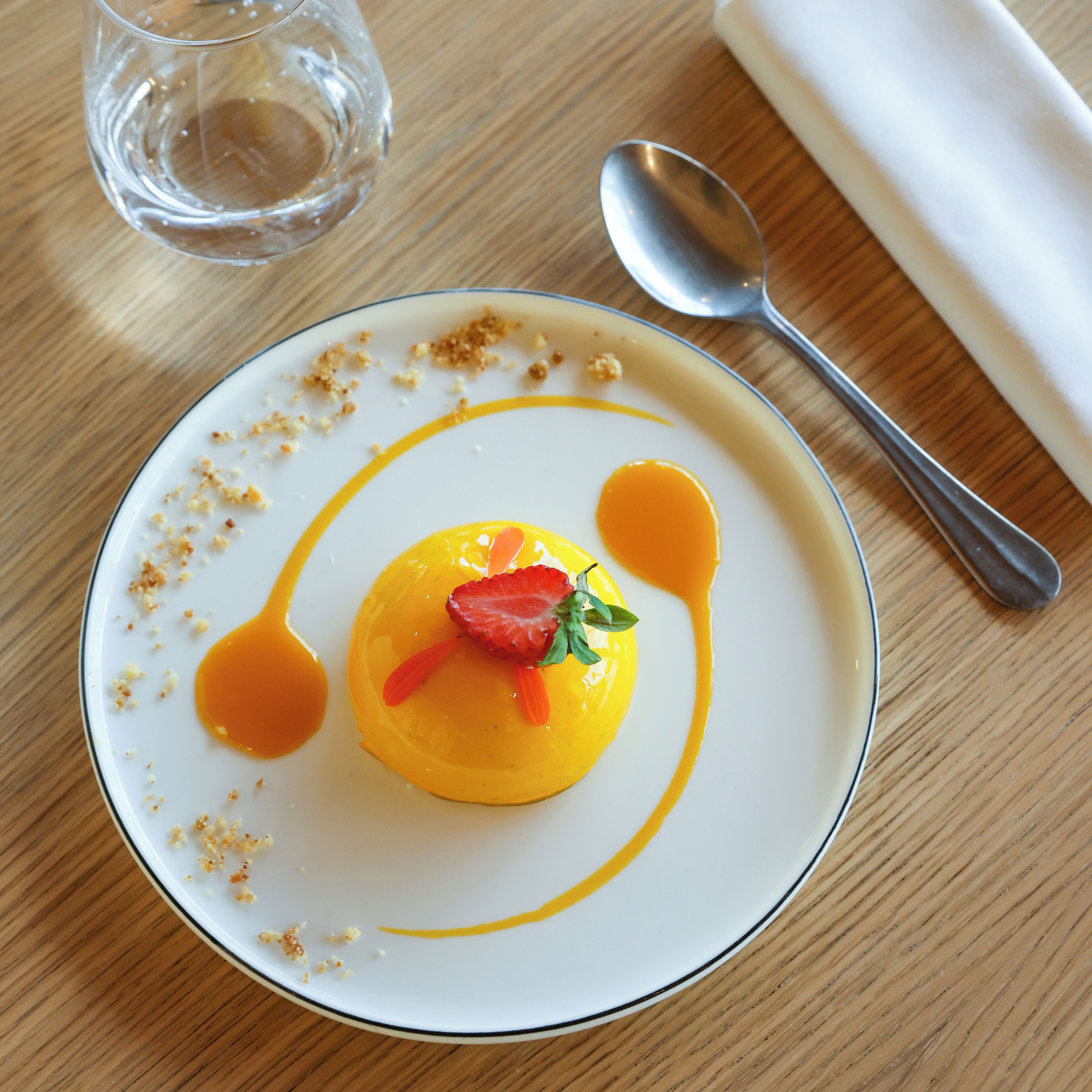 Un postre sobre un plato blanco, con una cuchara y un vaso de agua en una mesa de madera.