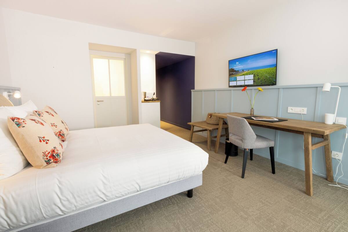 A hotel room with a bed, a desk, and a chair. There is a television on the wall and a window with a view of the outdoors.