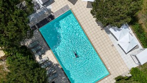 Vista aerea di una piscina rettangolare con una persona al centro, circondata da vialetti piastrellati e vegetazione.