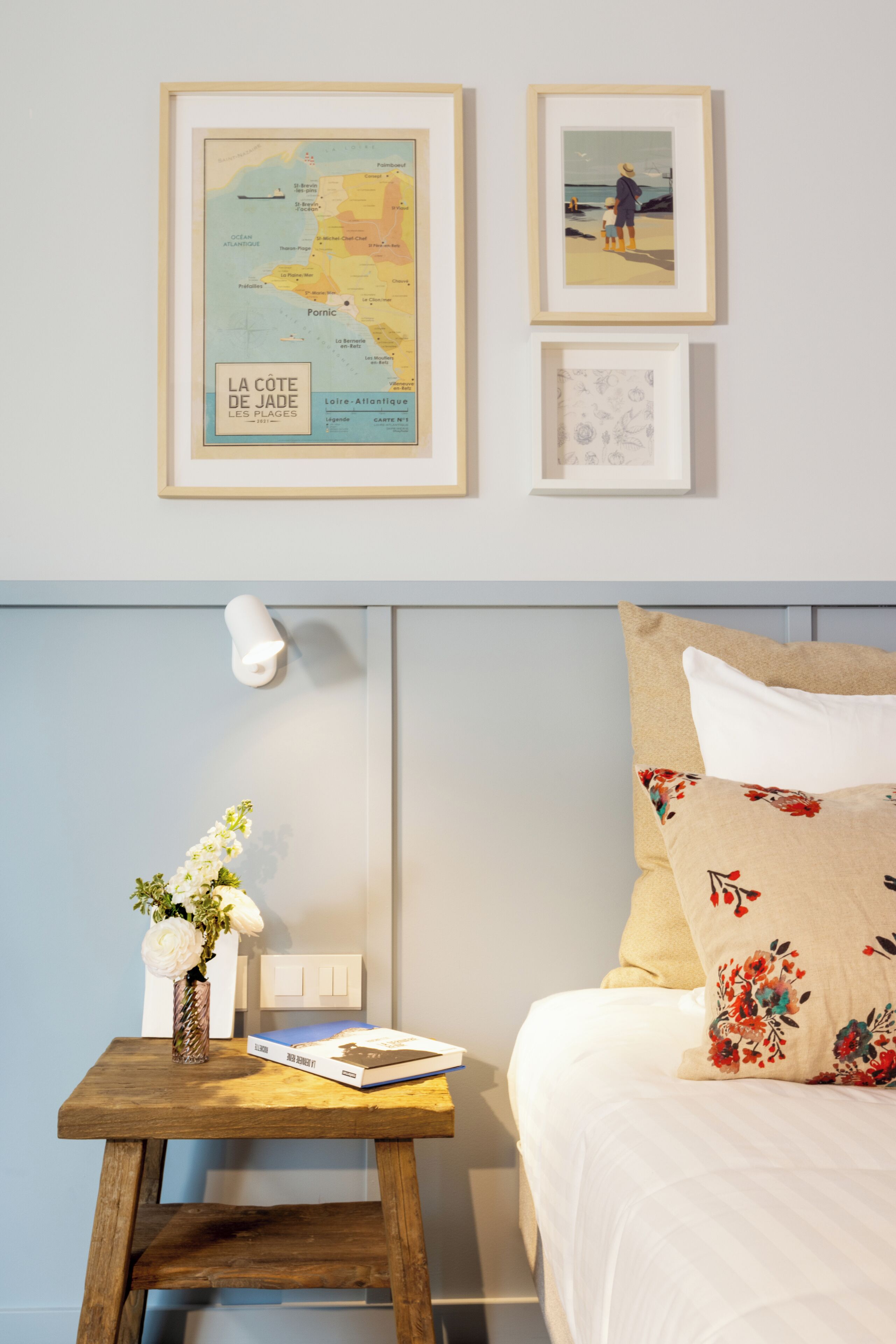 A neatly arranged bedroom with a bed, a bedside table with a book and a vase of flowers, a framed map, and a framed picture on the wall.