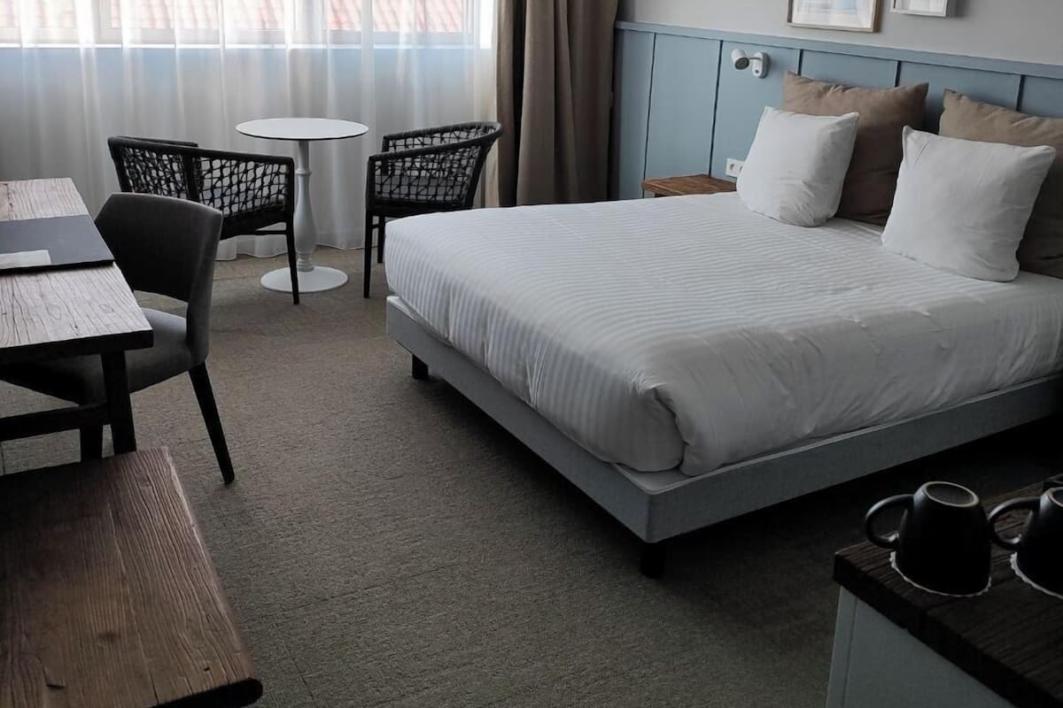 A hotel room with a bed, a desk, a chair, a small table, and a window with curtains.