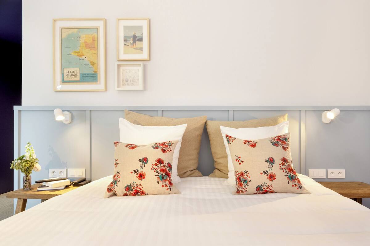 A neatly made bed with floral pillows, a bedside table with a book and a vase, and two framed pictures on the wall.
