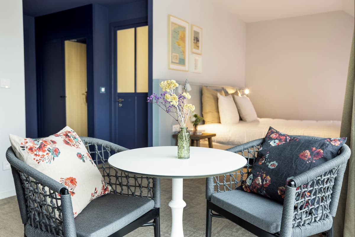 A room with two wicker chairs, a round white table, and a bed with floral pillows.
