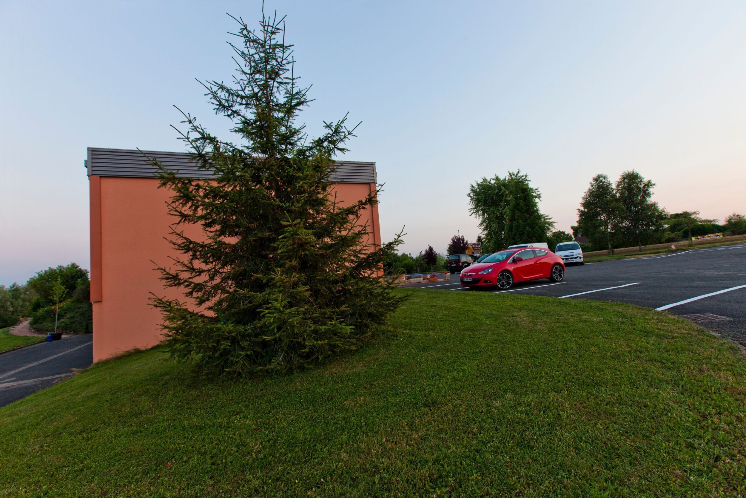 A building with a flat roof and a small tree in front. There is a red car parked on the street.