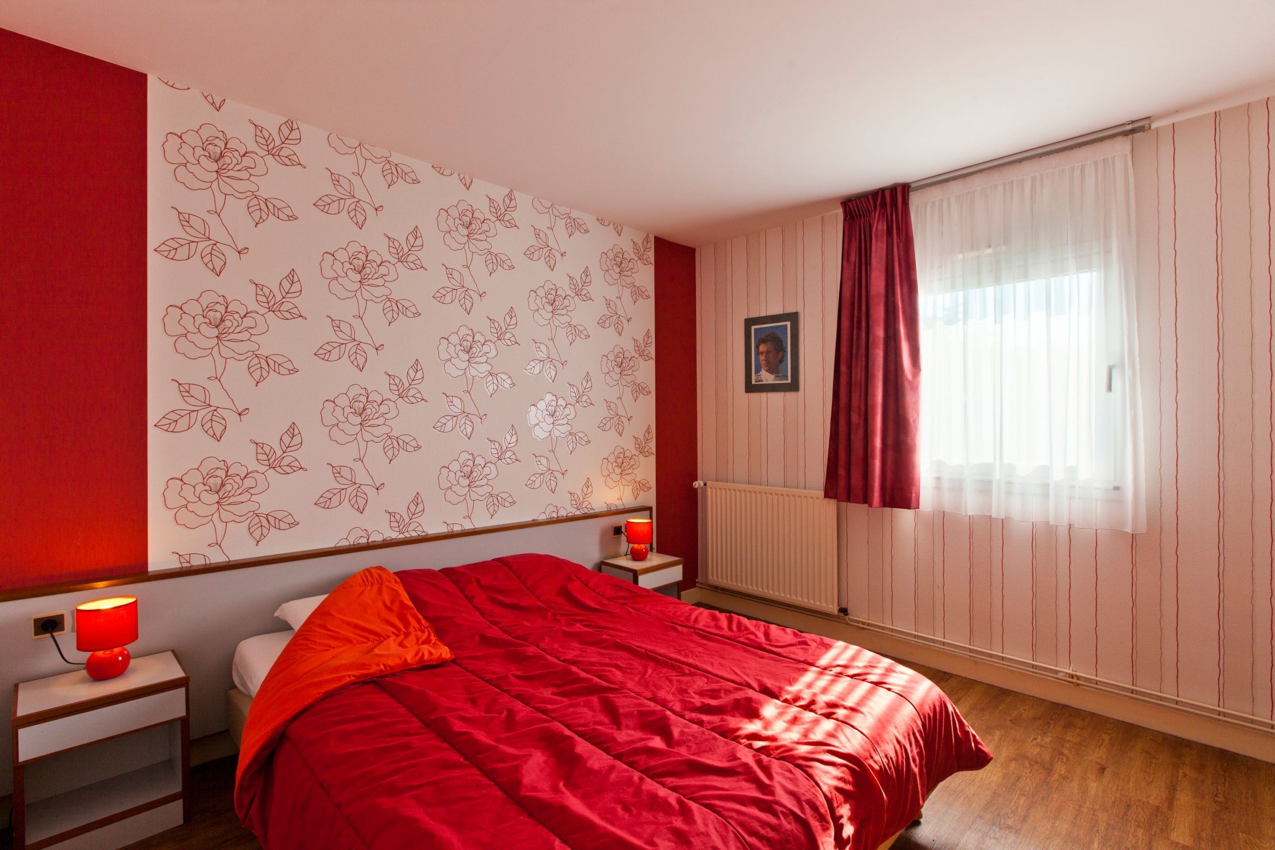 Un dormitorio con colcha roja, una mesita de noche con lámpara, un cuadro enmarcado en la pared y una ventana con cortinas.
