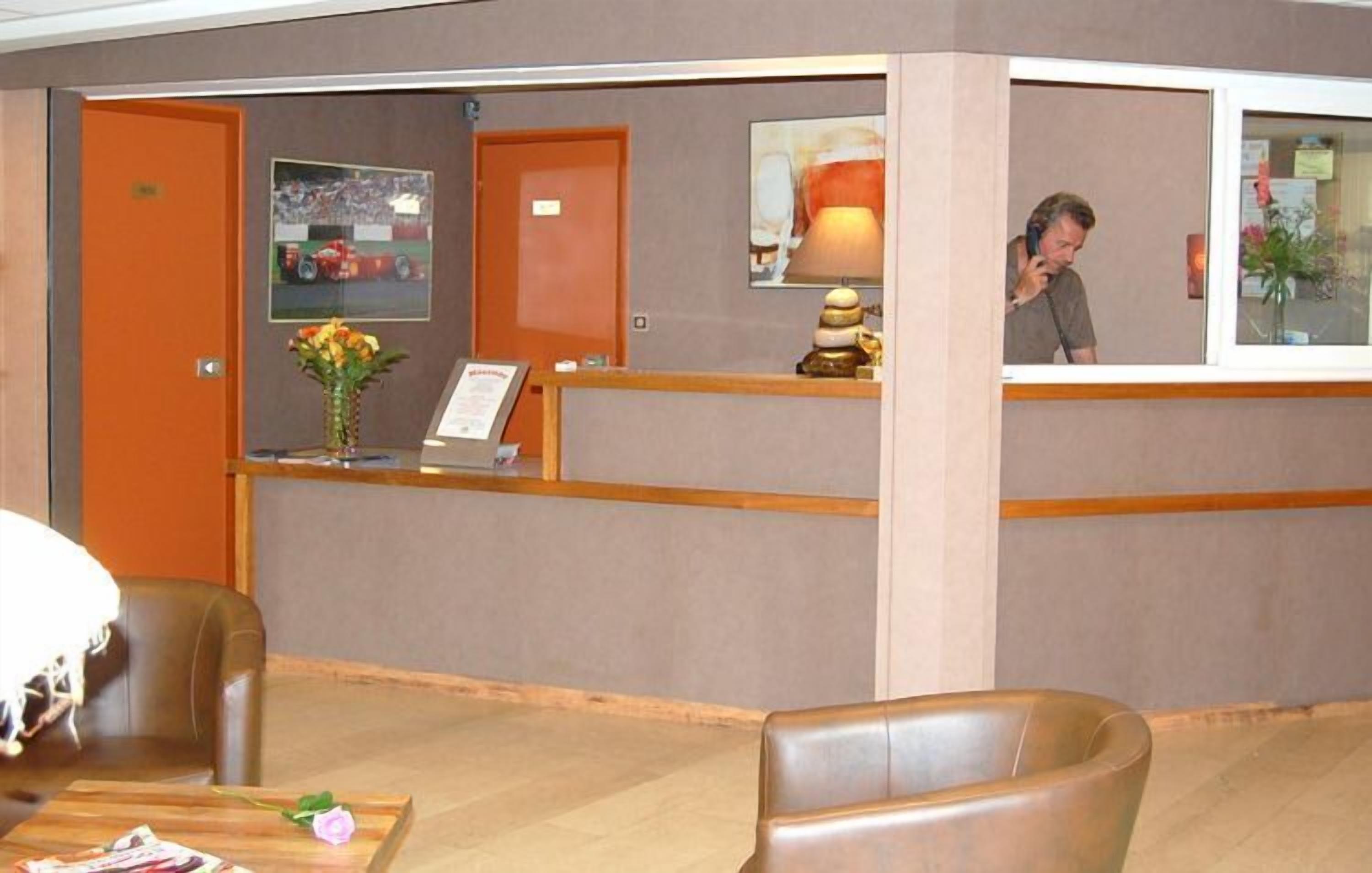 A hotel reception area with a reception desk, a framed picture on the wall, a vase with flowers, and a lamp.