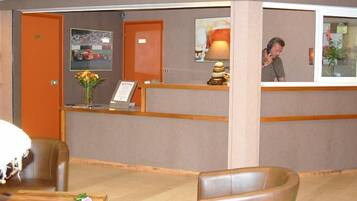 A hotel reception area with a reception desk, a framed picture on the wall, a vase with flowers, and a lamp.