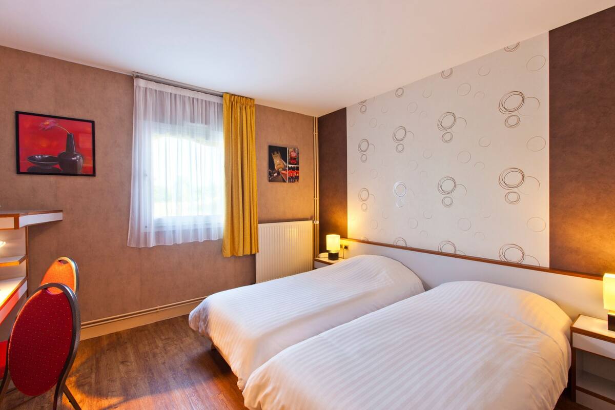 A hotel room with two beds, a desk, a chair, a window with curtains, and wall art.