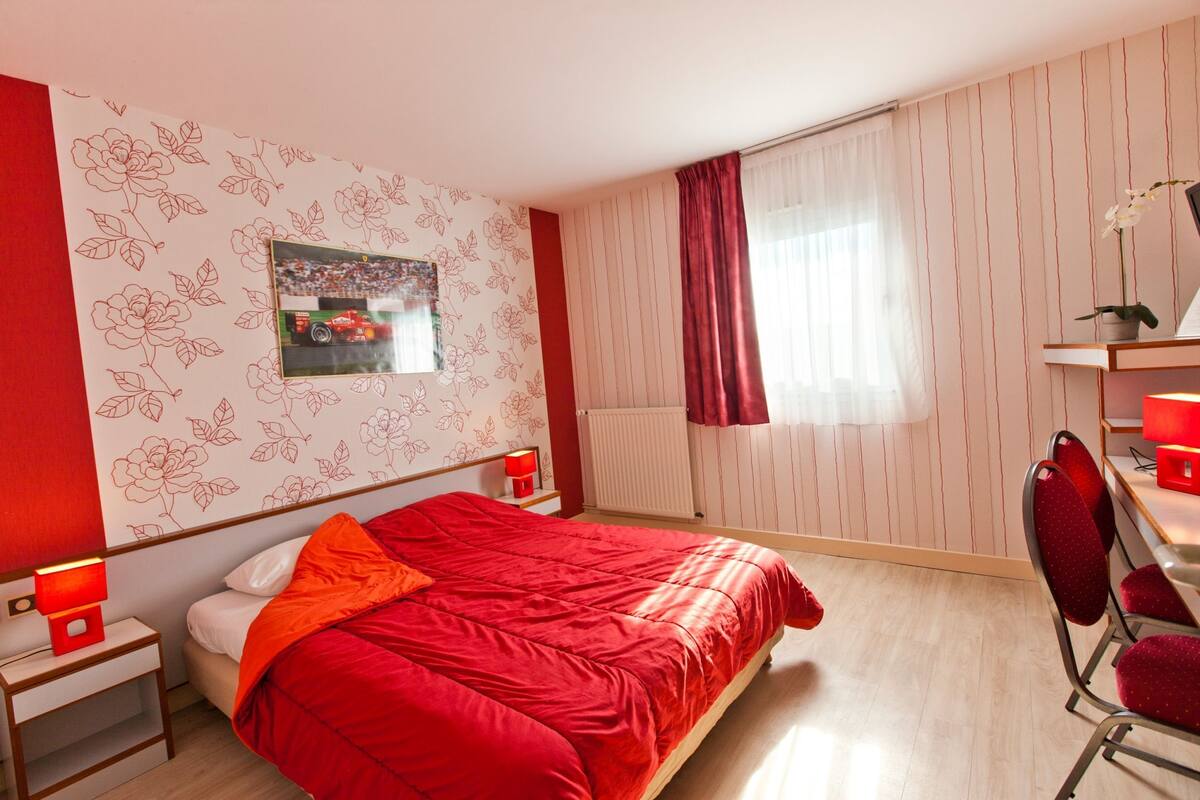 A bedroom with a bed, a desk, a chair, a nightstand, a lamp, a window with curtains, and a wall with floral wallpaper.