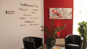 Et receptionsområde med en rød væg, en velkomstskilt på flere sprog, to sorte stole, et bord med en menu og en potteplante.