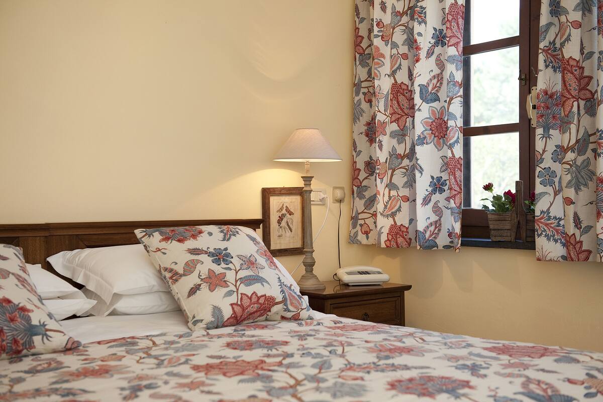 A bedroom with floral bedding, a bedside lamp, a nightstand with a phone, and curtains with a floral pattern.