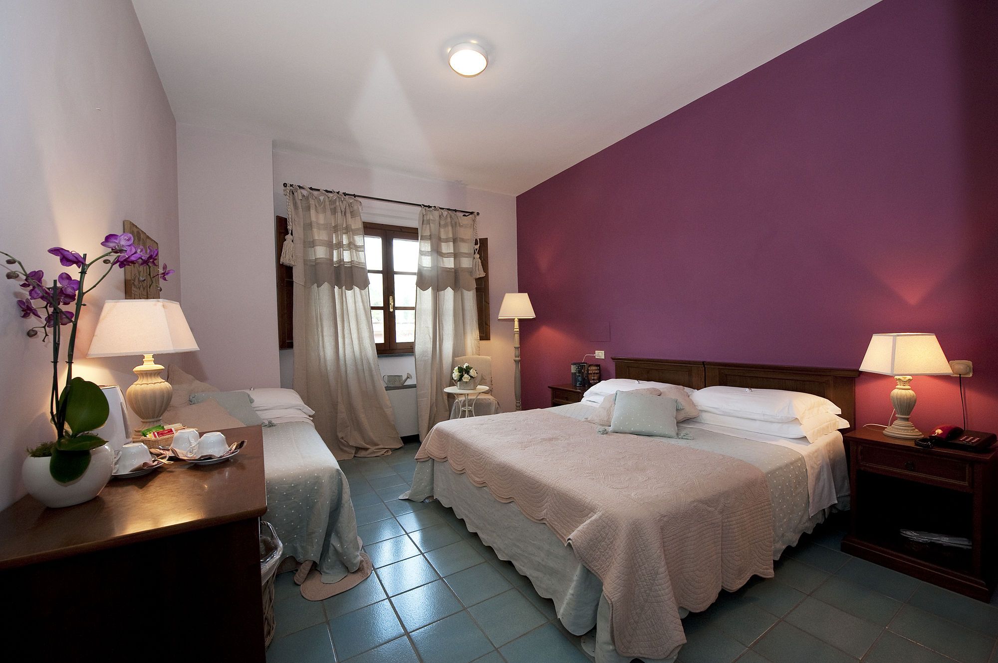 A hotel room with two beds, a window with curtains, a bedside table with a lamp, and a small table with a plant and cups.
