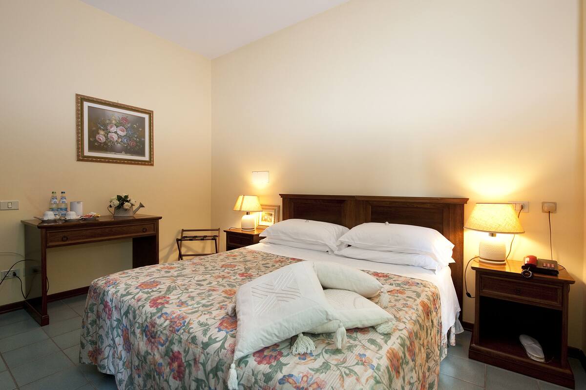 A neatly made bed with floral bedding, two bedside lamps, a wooden nightstand, and a desk with a lamp and a vase of flowers.