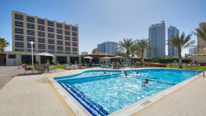 A swimming pool with people swimming and sunbathing, surrounded by palm trees and buildings. - Ajman Beach Hotel (Ajman)