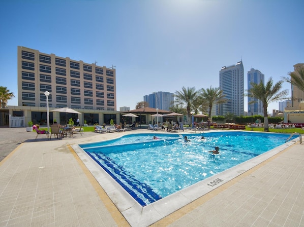 A swimming pool with people swimming and sunbathing, surrounded by palm trees and buildings.