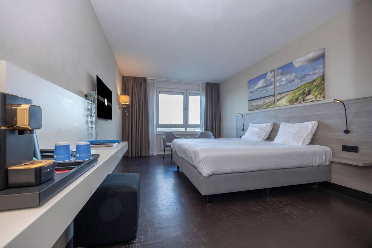 Quarto de hotel com uma cama, uma escrivaninha com cafeteira, televisão e uma janela com cortinas.
