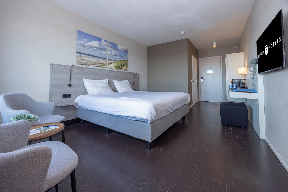 Quarto de hotel moderno com uma cama grande, uma escrivaninha com luminária, uma cadeira e uma televisão na parede.