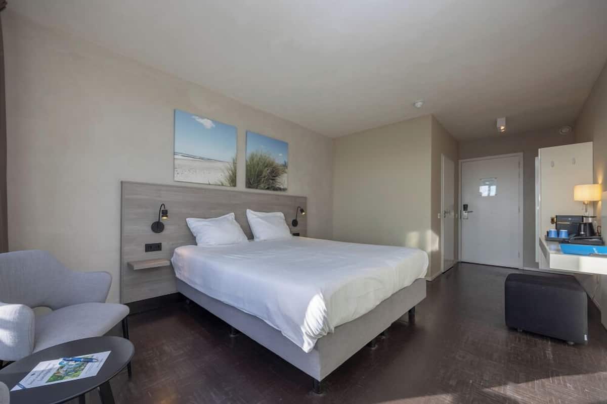 Quarto de hotel com uma cama, uma escrivaninha, uma cadeira e uma mesa pequena.